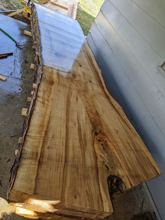 Live-edge slab of light-colored wood, possibly a tabletop, rests against a white wall and concrete.