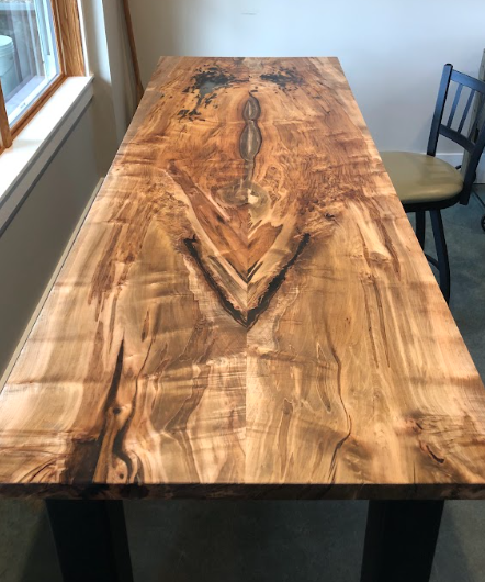Wooden table with unique grain pattern, dark legs, near a window and chair.