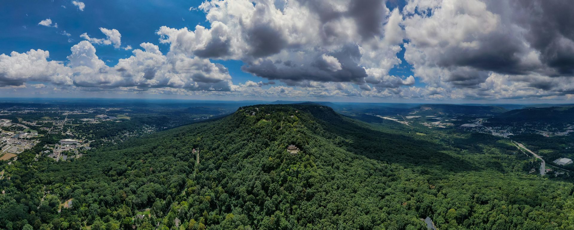 Overhead shot of a forested mountain under a partly cloudy blue sky. In the distance is a city.