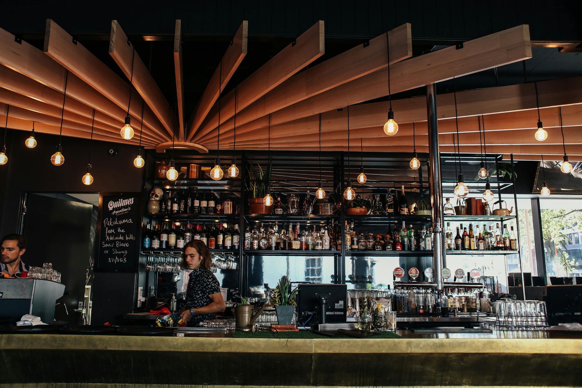 Bar with bartender, bottles of alcohol, wooden ceiling detail, and hanging lights.