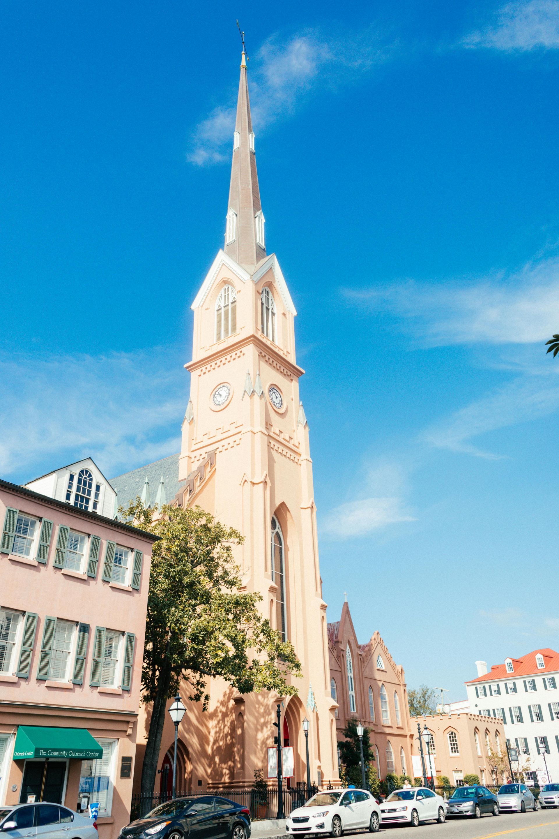 Tall, light-colored church with a steeple against a bright blue sky. Cars are parked along the street in front of it.