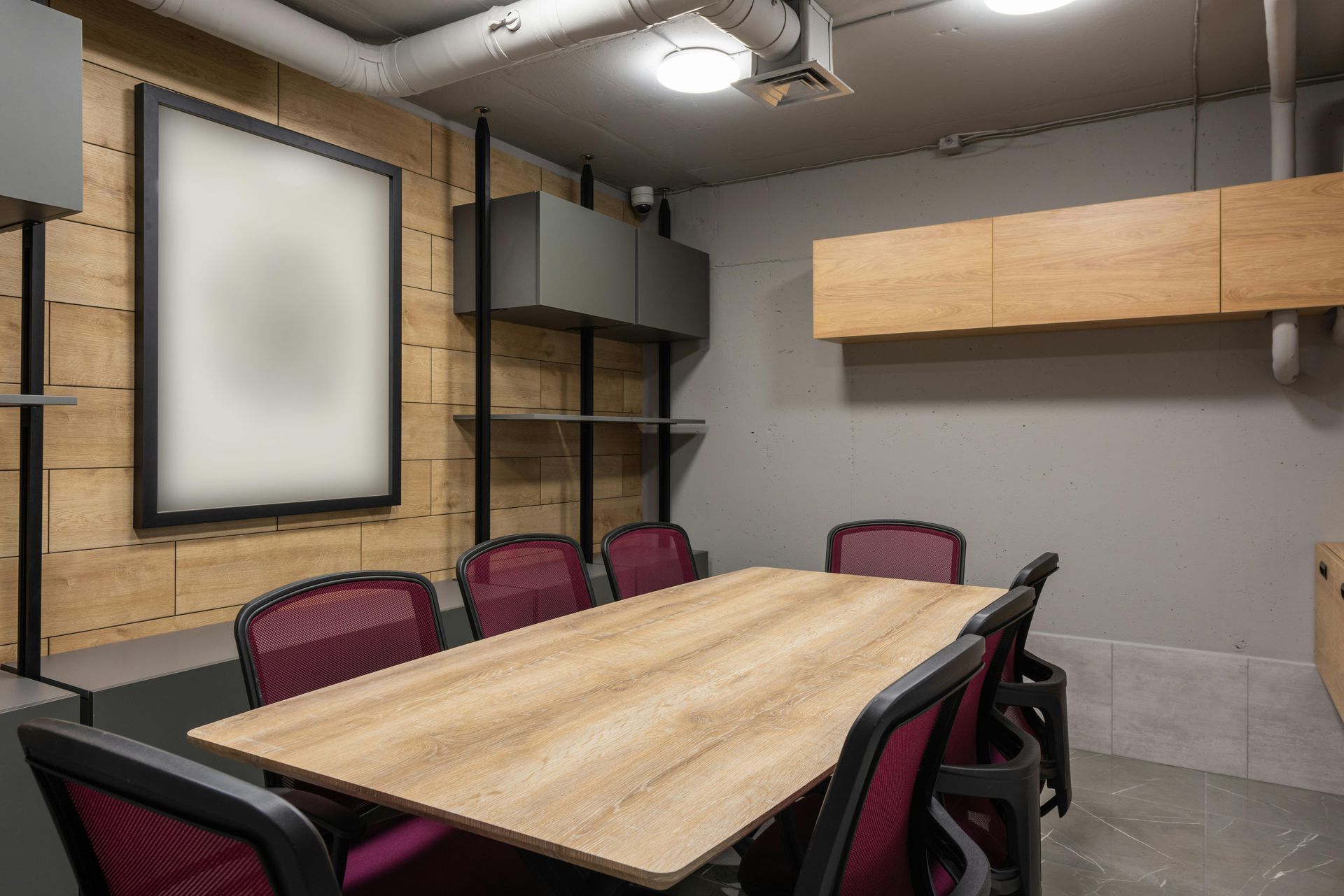 Conference room with a long wooden table, red chairs, shelves, and a wall-mounted screen