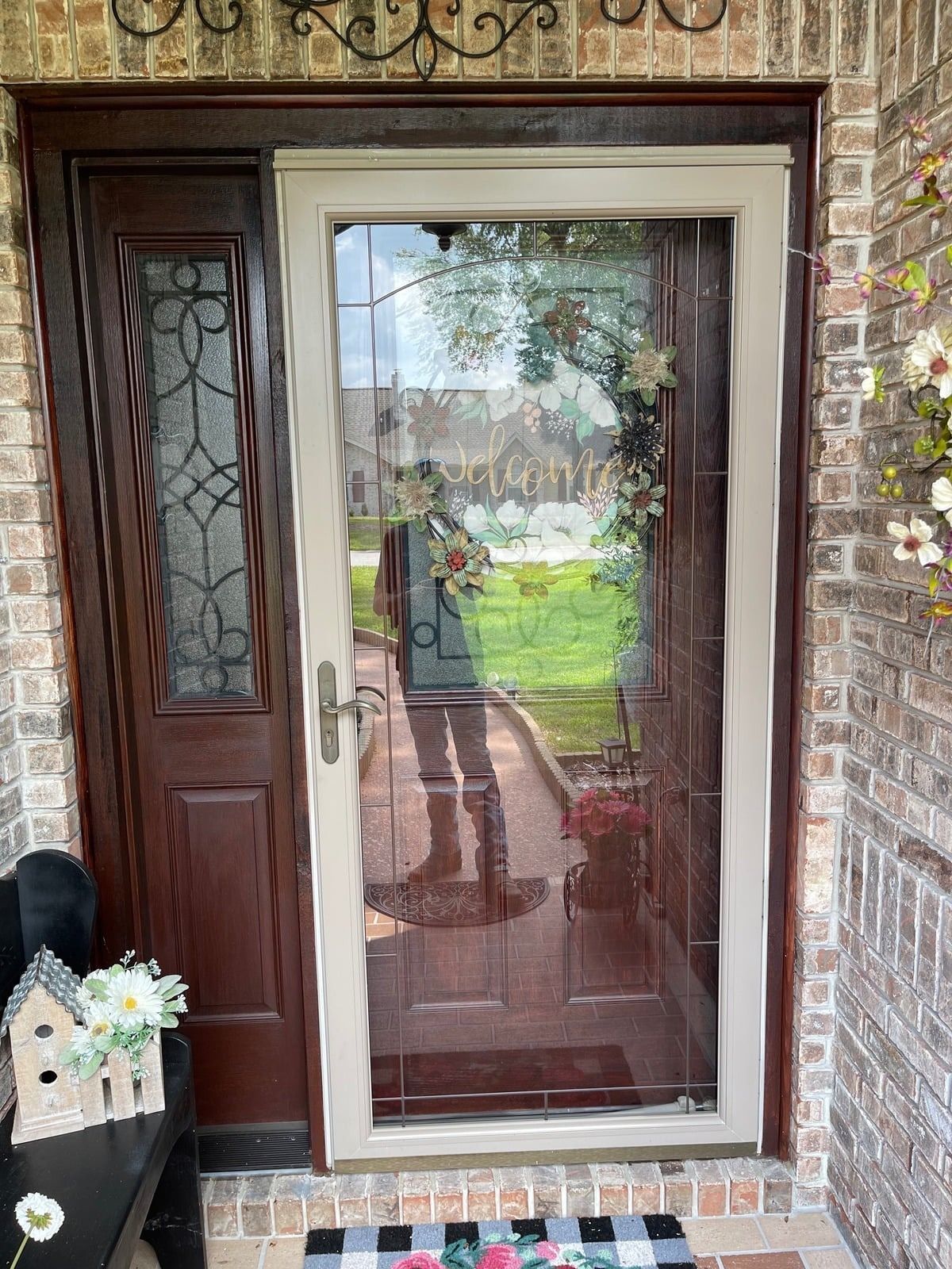 Front door with glass panel and side window set in a brick entryway, reflecting a garden and person.