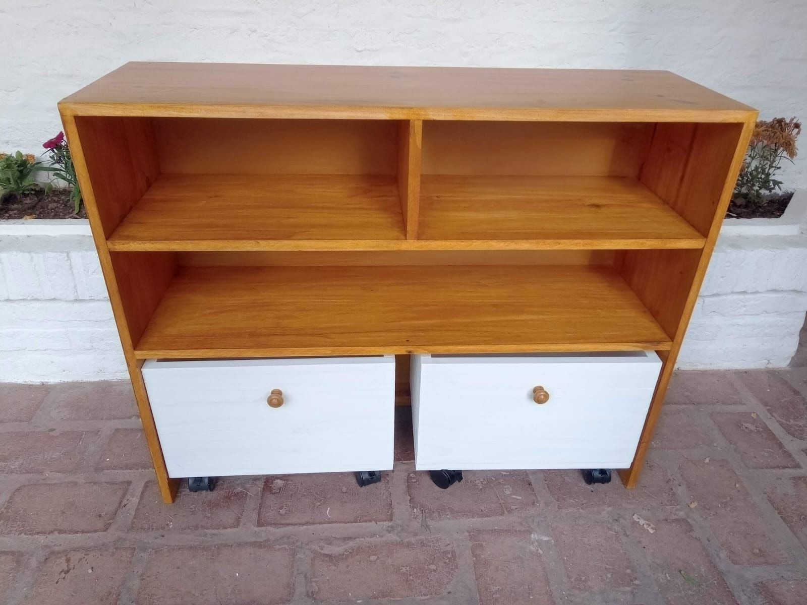 Estantería de madera con dos estantes abiertos, dos cajones blancos y ruedas.