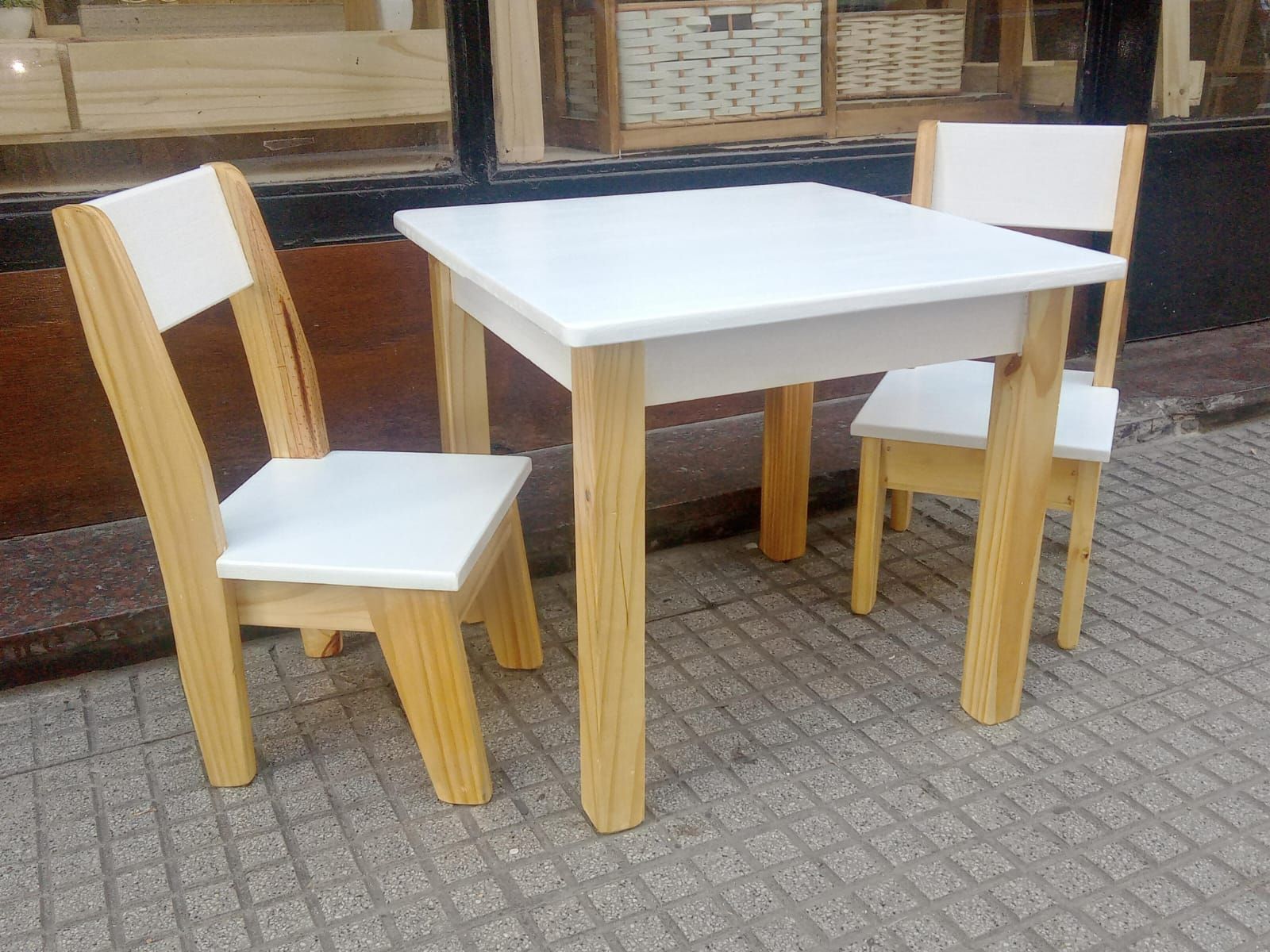 Mesa infantil cuadrada blanca y dos sillas en una acera. Los asientos de la mesa y las sillas son blancos; las patas son de madera clara.