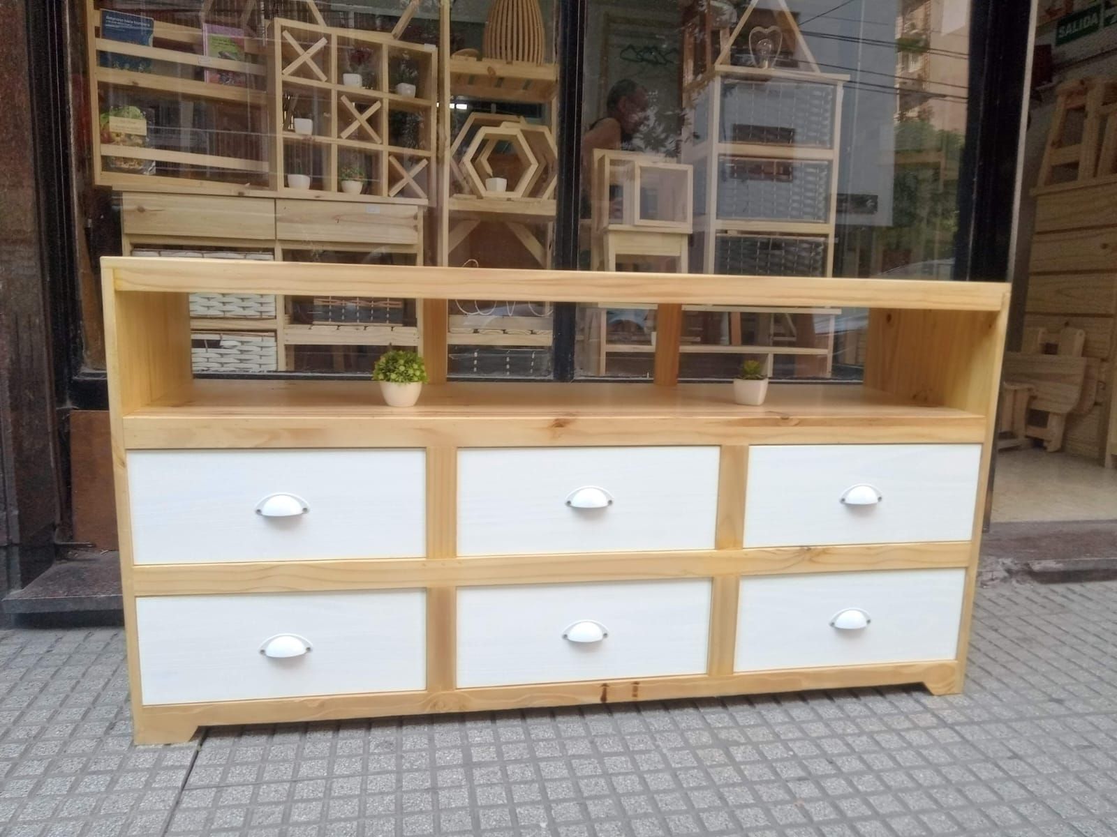 Mueble de TV de madera con seis cajones blancos y estantes abiertos, expuesto en el exterior de una tienda.