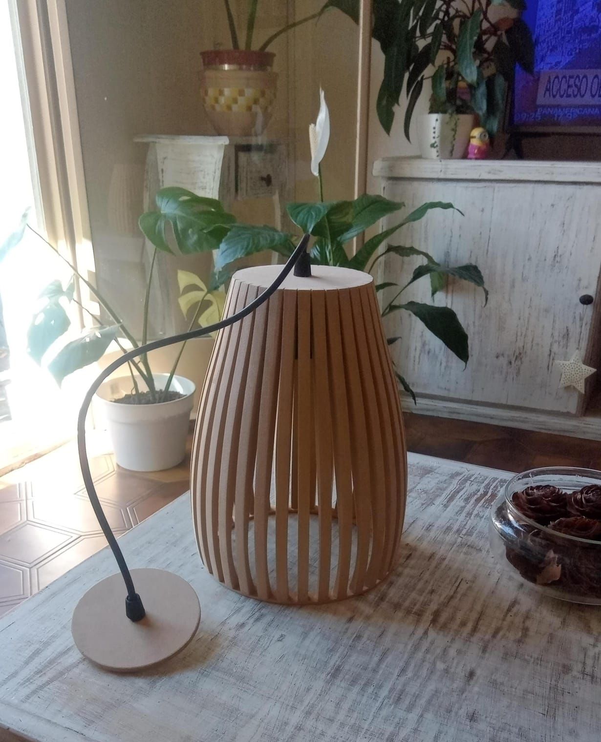 Lámpara de madera con cuello flexible, colocada sobre una alfombra con plantas de fondo.