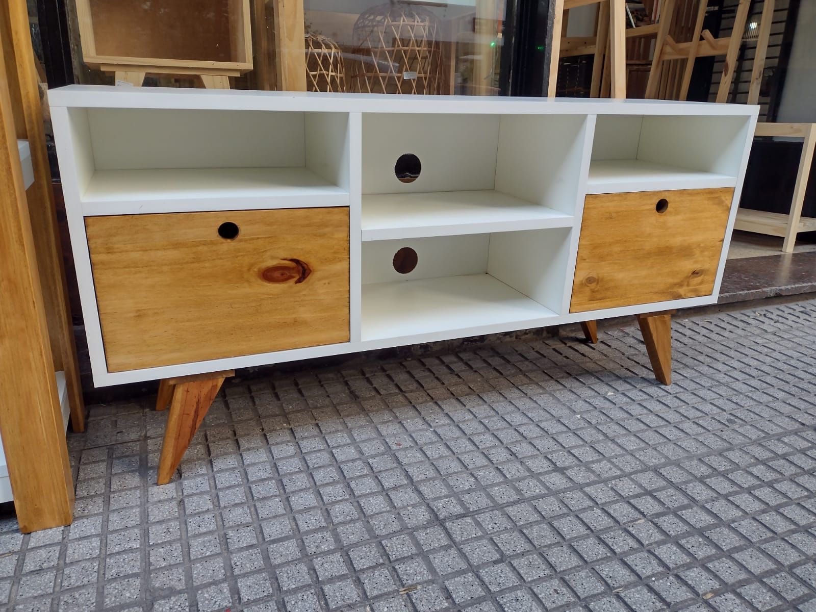 Mueble de TV en color blanco y madera con dos cajones, estantes abiertos y patas en ángulo.