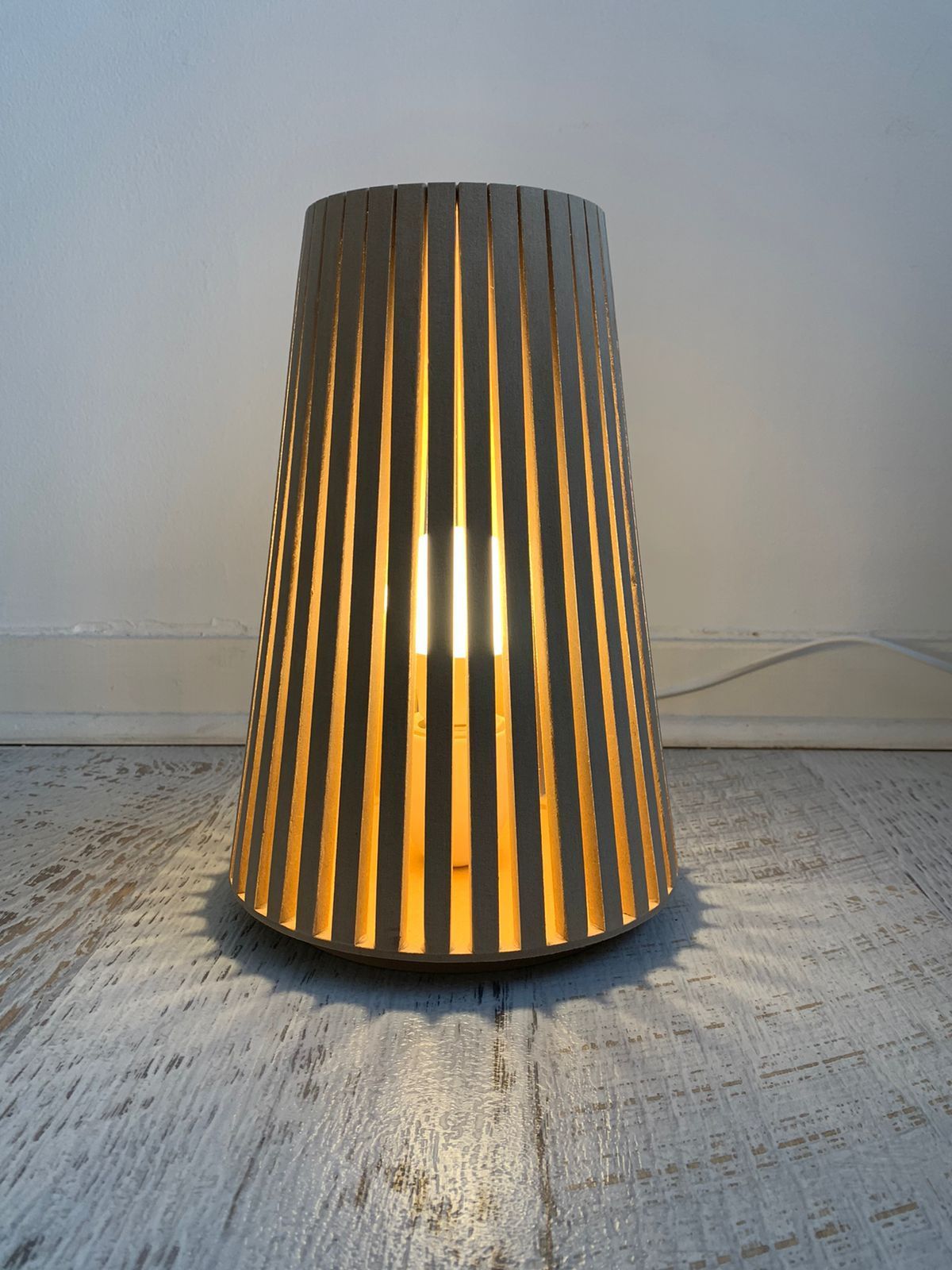 Lámpara de madera con ranuras verticales que proyectan luz y sombra sobre el suelo y la pared.