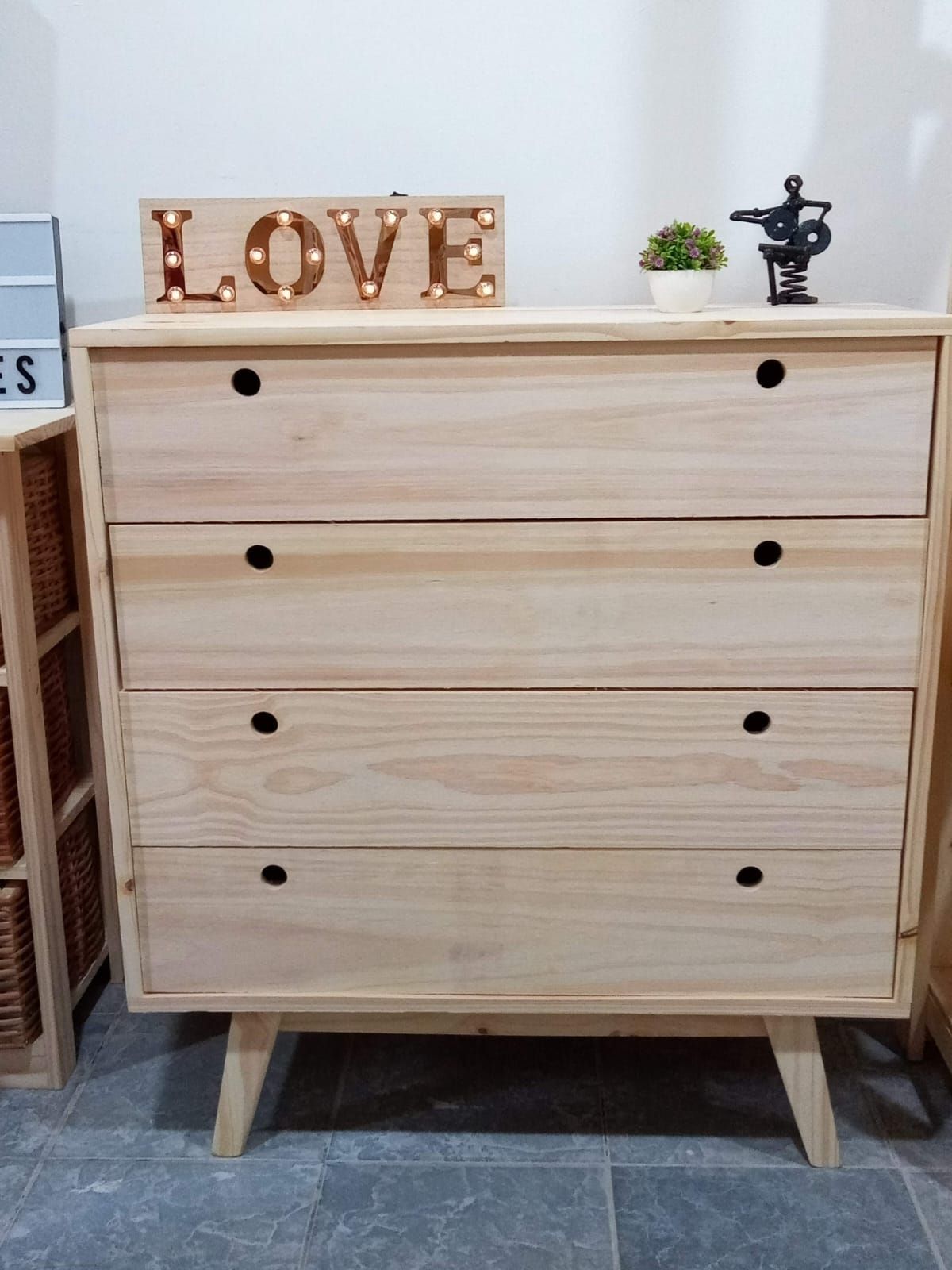 Cómoda de madera de cuatro cajones con pequeños tiradores negros, patas en ángulo y elementos en la parte superior.