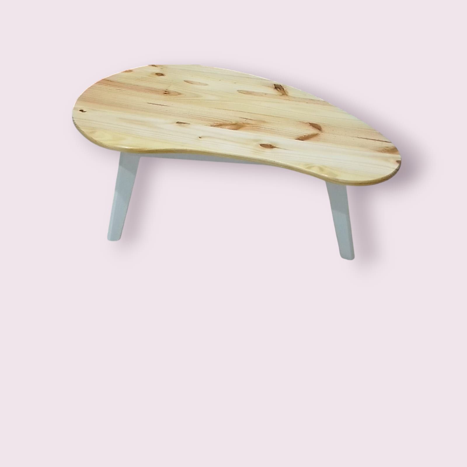 Mesa de centro en forma de lágrima, de color madera clara, con patas blancas sobre un fondo rosa.