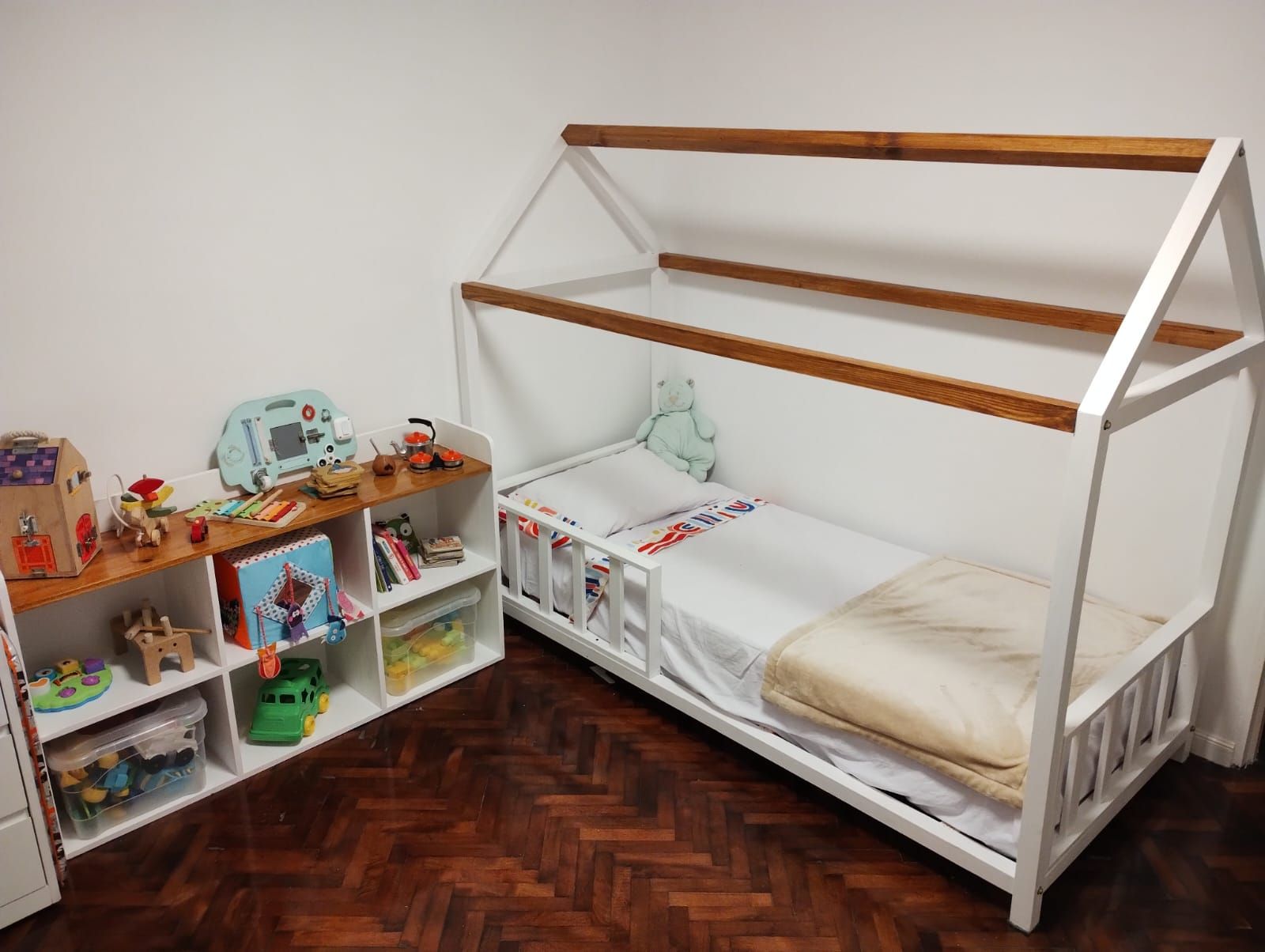 Habitación infantil con cama blanca en forma de casa y estante de madera lleno de juguetes, contra una pared blanca.