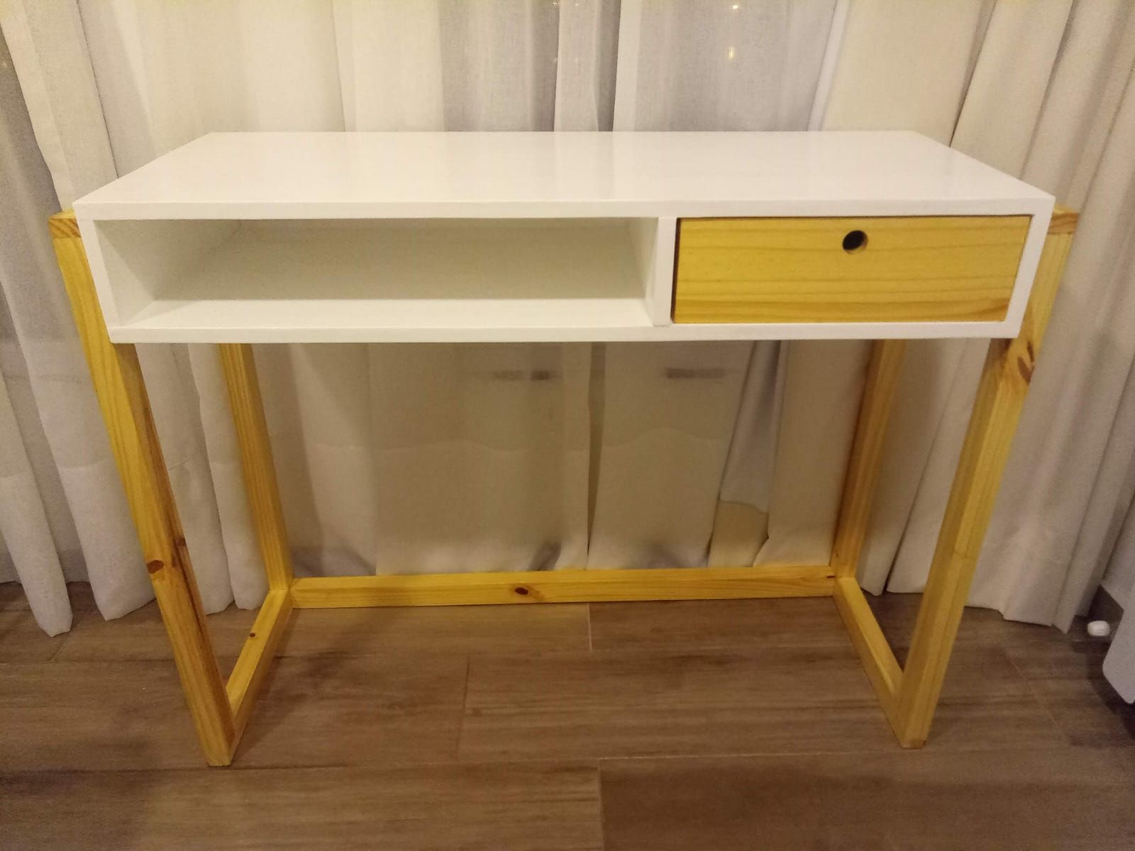 Escritorio blanco y de madera con cajón y estante abierto, sobre un fondo de cortina blanca.