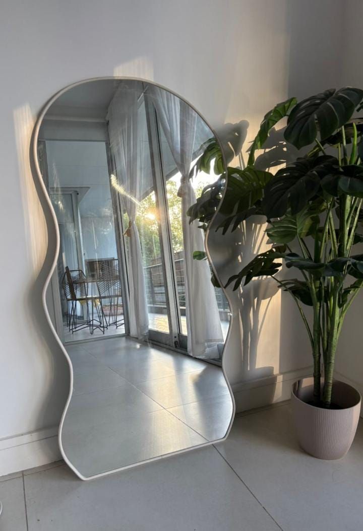Espejo de borde ondulado apoyado sobre una pared blanca, que refleja una habitación con luz natural y una planta en maceta.
