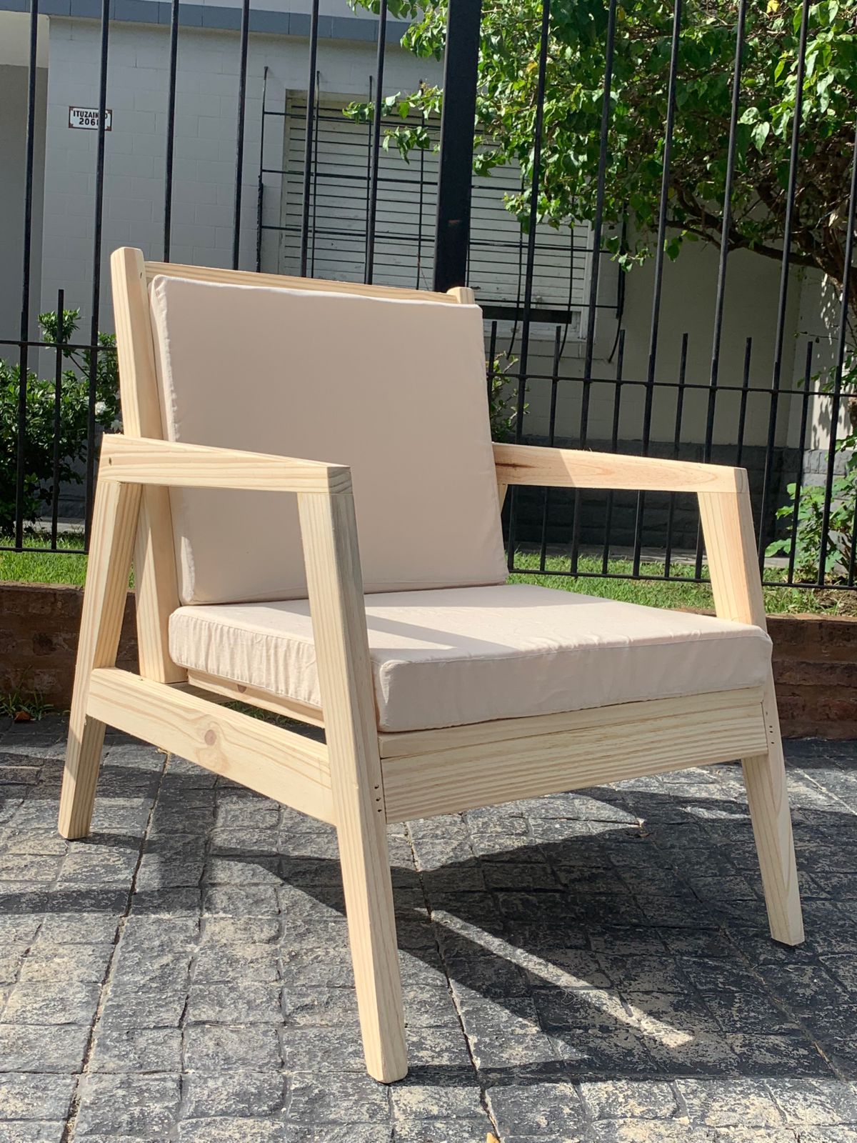 Sillón de madera con cojines beige en un patio de ladrillo, contra una valla y follaje.
