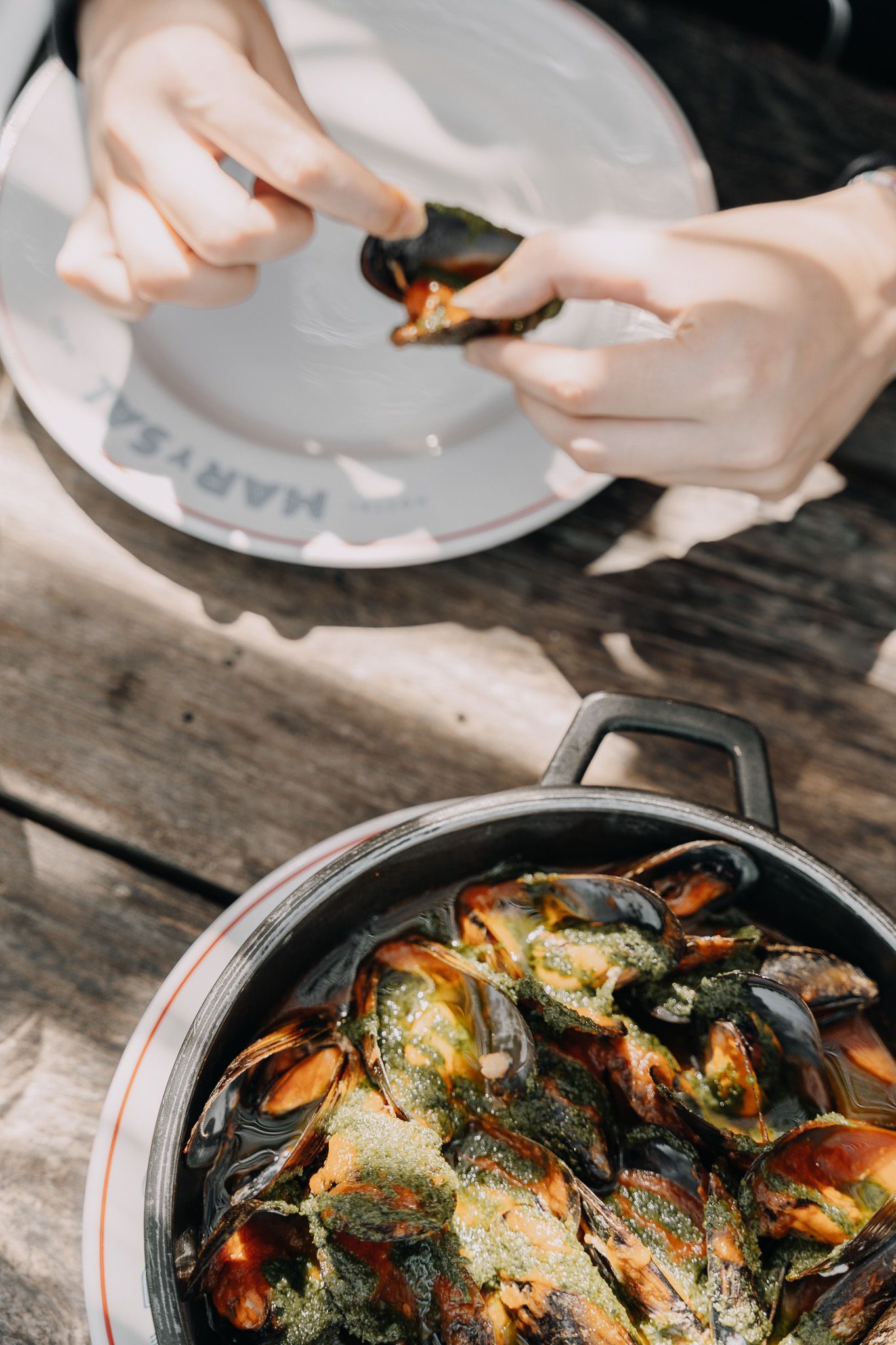 Unas manos sostienen una concha de mejillón abierta sobre un plato, mientras que una sartén de hierro fundido llena de mejillones cocidos reposa sobre una mesa de madera.