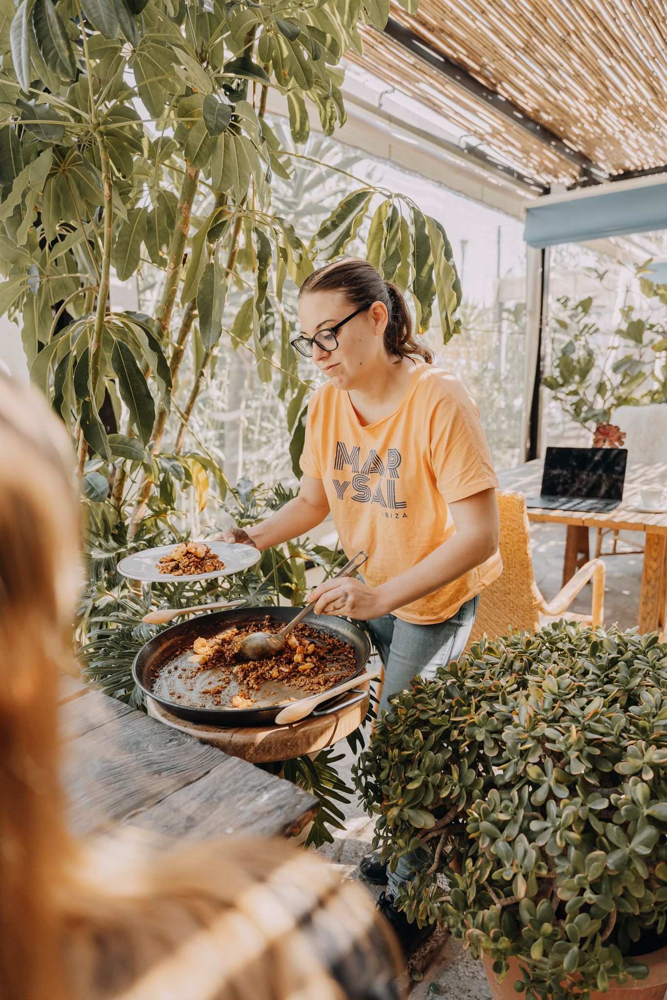 Una persona con una camisa color melocotón sirve comida de una sartén grande en un plato blanco en un ambiente interior soleado y lleno de plantas.