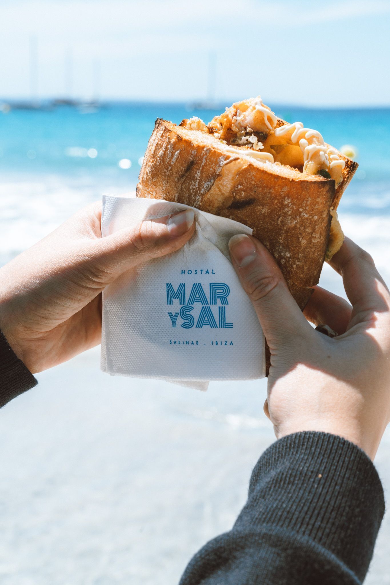 Unas manos sostienen un sándwich envuelto en una servilleta con el logo de la marca, con el azul brillante de la playa como telón de fondo.