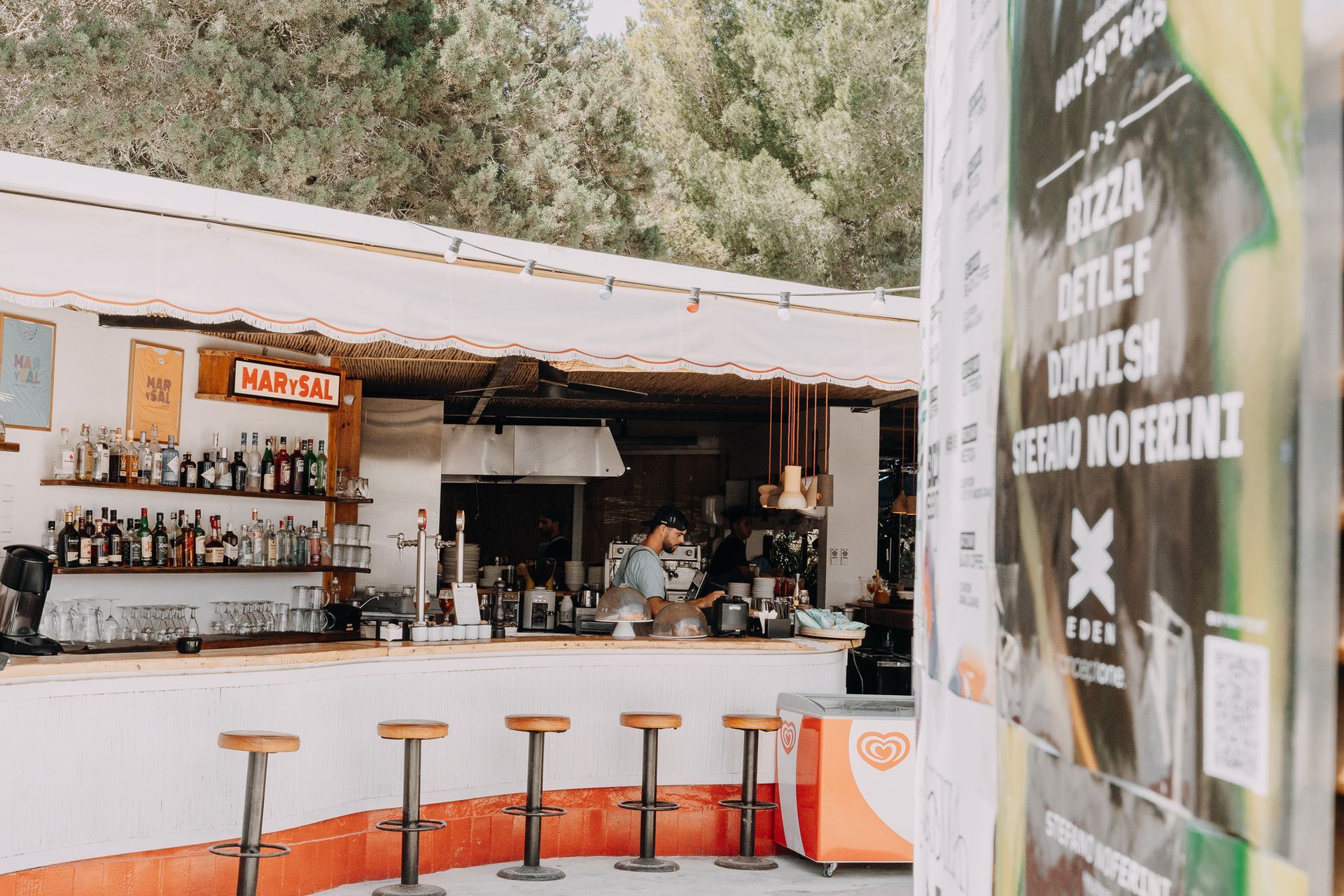 Un bar al aire libre con una larga barra blanca, taburetes y estantes con botellas de licor, visto junto a un cartel de un concierto.