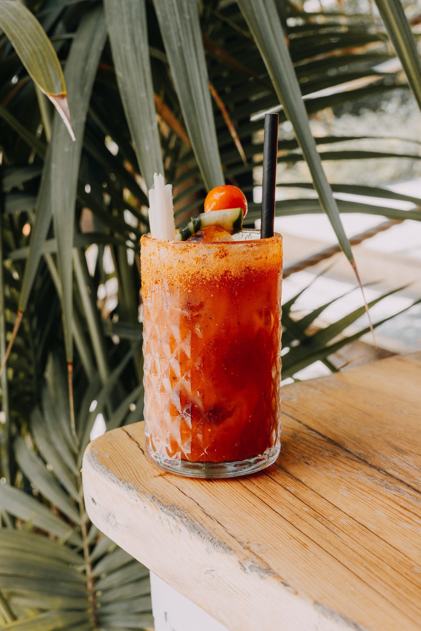 Un vaso texturizado con un cóctel rojo, borde especiado, pajita negra y guarnición de tomate, sobre una mesa de madera.