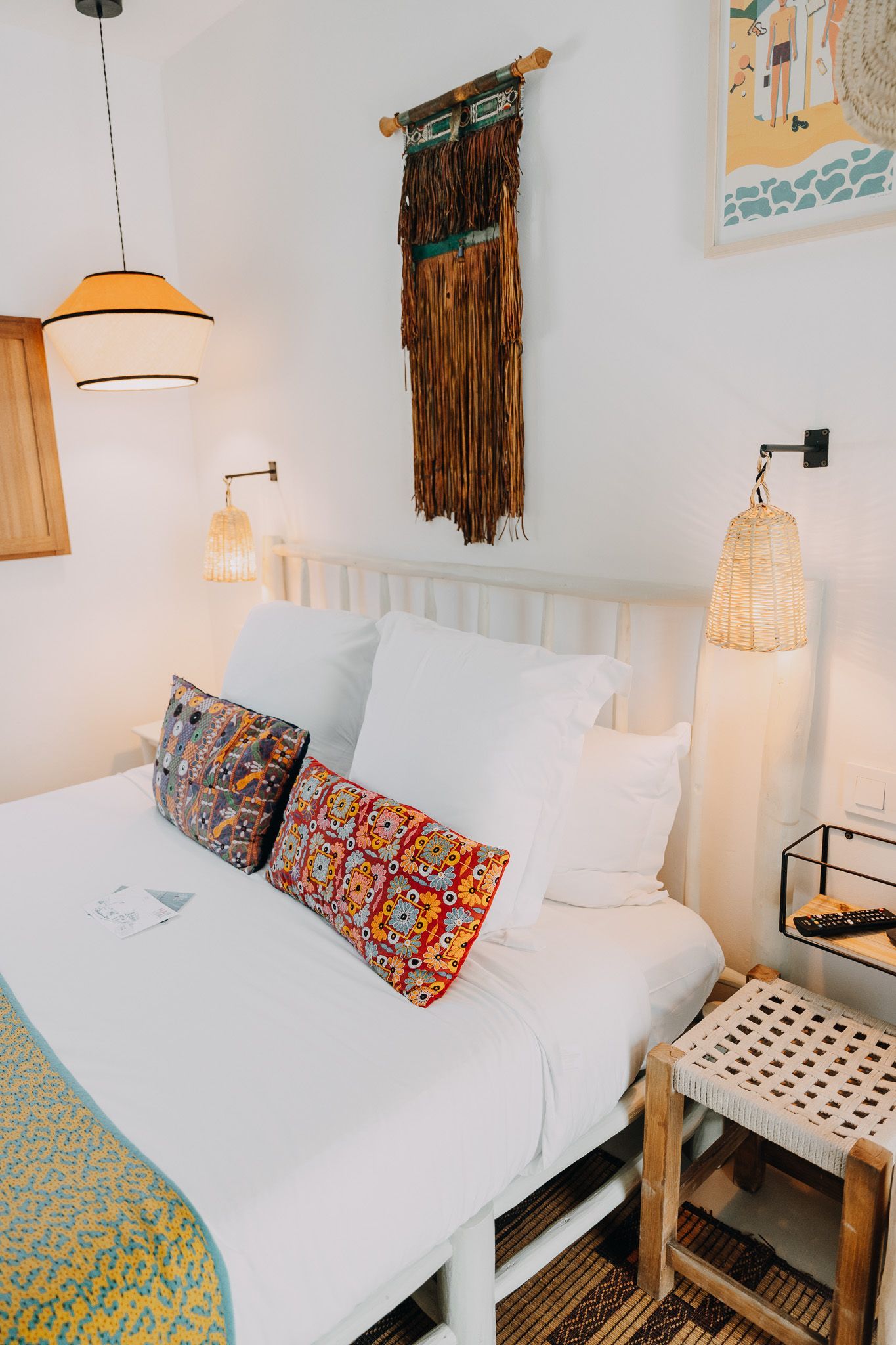 Un dormitorio acogedor cuenta con una cama con sábanas blancas, cojines decorativos estampados, un tapiz en la pared y lámparas de noche tejidas.
