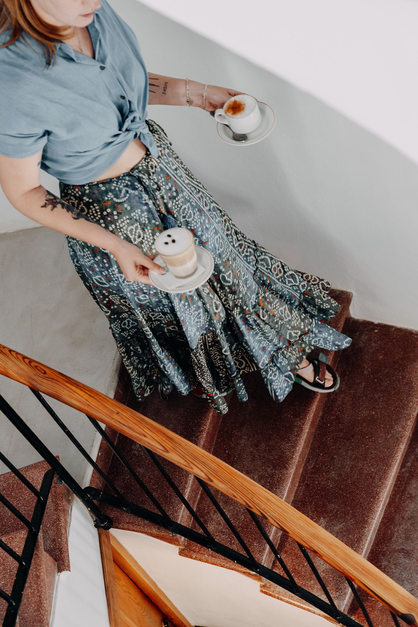 Una persona con una camisa azul y una falda estampada baja unas escaleras sosteniendo dos tazas de café sobre sus platillos.
