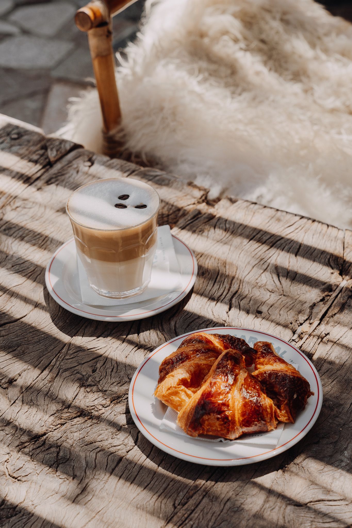 Un café con leche en un vaso y un pastel en un plato reposan sobre una mesa de madera desgastada bajo la luz del sol a rayas.