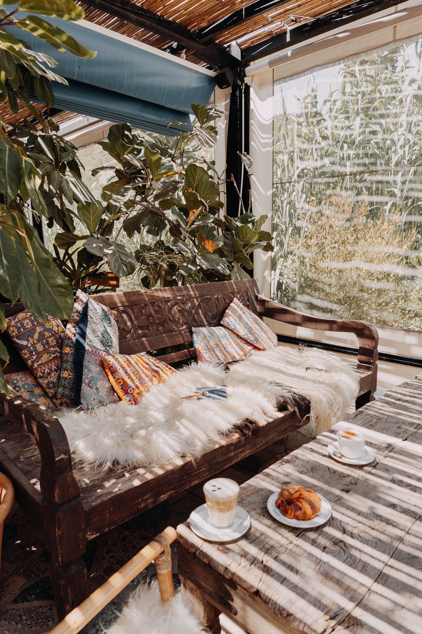 Acogedora terraza con un sofá de madera oscura, mantas de piel de oveja y una mesa puesta con café y un cruasán.