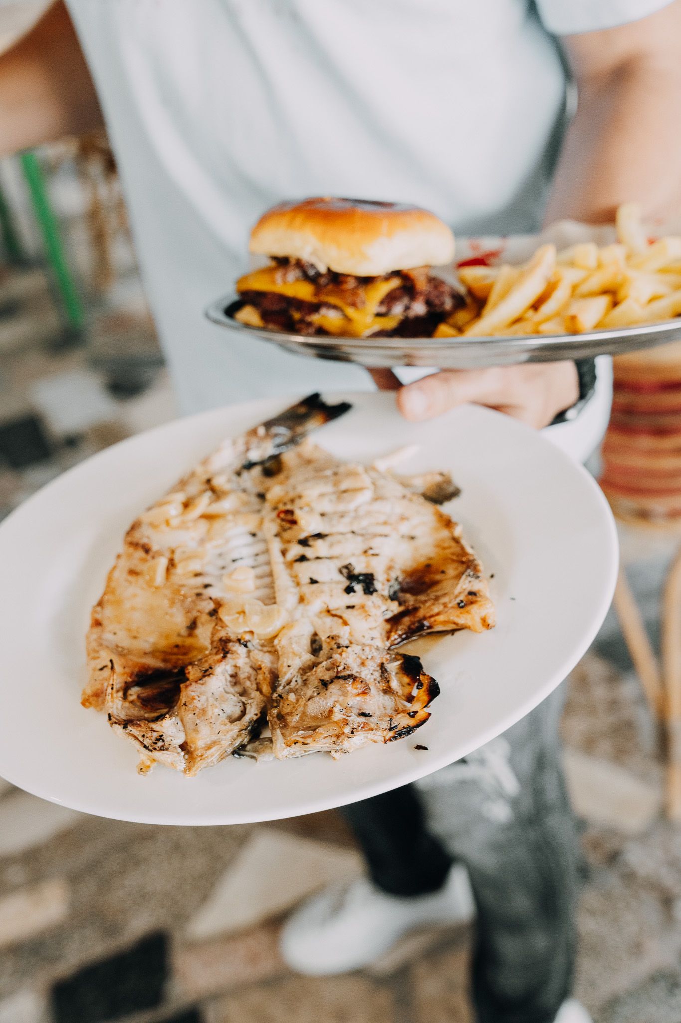 Una persona sostiene dos platos: uno con un pescado entero a la parrilla y otro con una hamburguesa con queso y patatas fritas.