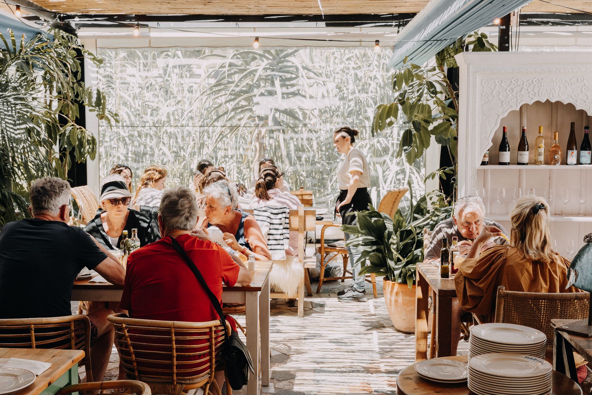 Un grupo de personas cenando en mesas en un luminoso patio interior lleno de plantas y con muebles de colores claros.
