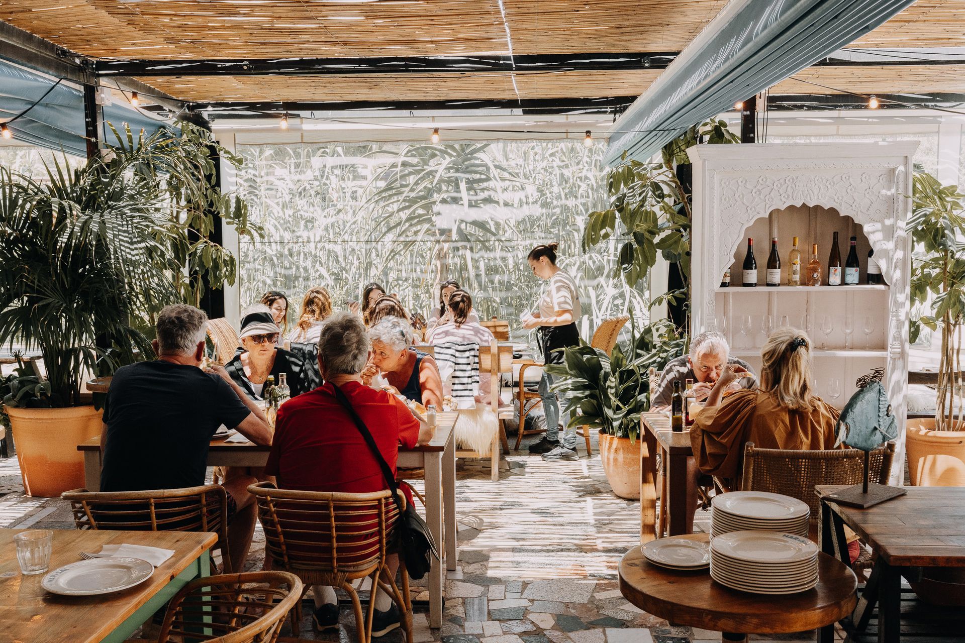 Gente cenando en un restaurante al aire libre con plantas exuberantes, muebles de madera y una estantería blanca al fondo.