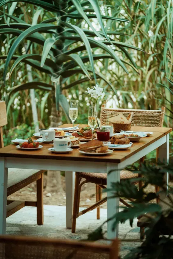 A table with plates of food and wine glasses on it is surrounded by plants.