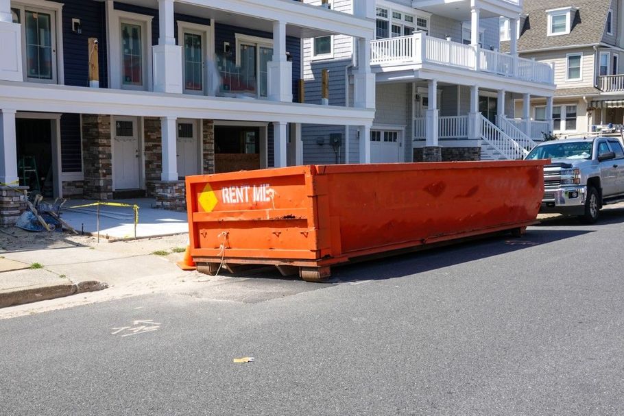 Orange construction dumpster on a residential street beside houses and parked cars.