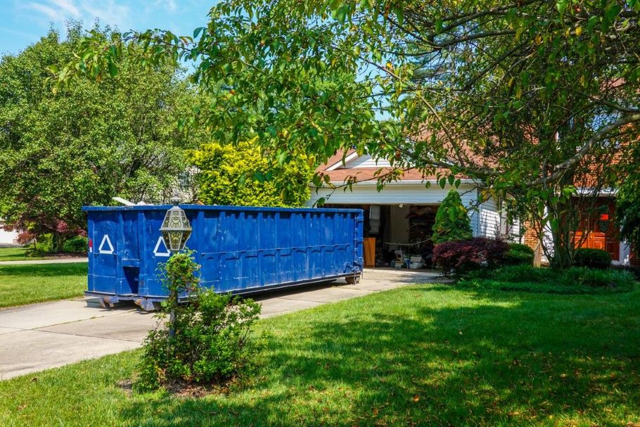 Large blue dumpster in a driveway beside a house, surrounded by green trees and lawn.
