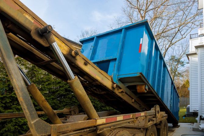 Blue dumpster being lifted by a refuse truck conveyor near a building outdoors