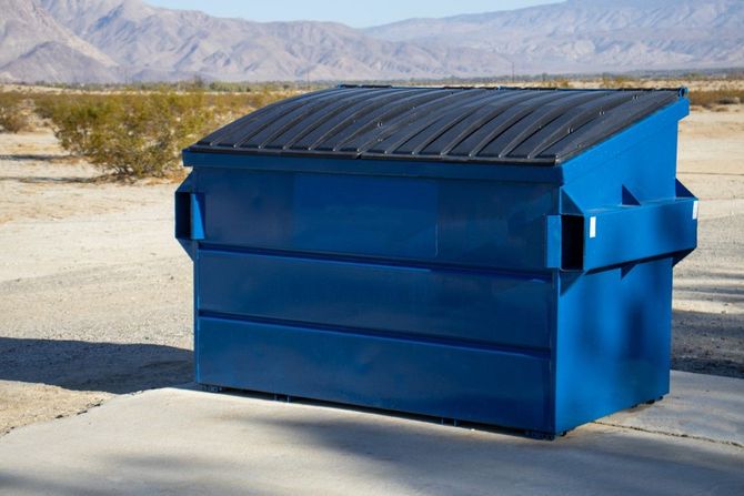 Blue dumpster with black lid on a concrete pad in a desert area with mountains in the background