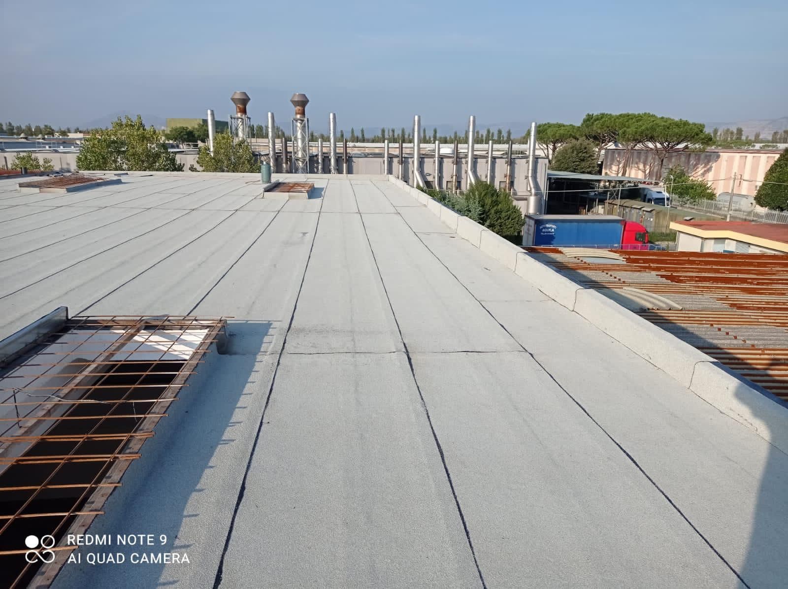 Tetto in costruzione con membrana bianca, tubi di ventilazione e, in lontananza, edifici industriali sotto un cielo azzurro.