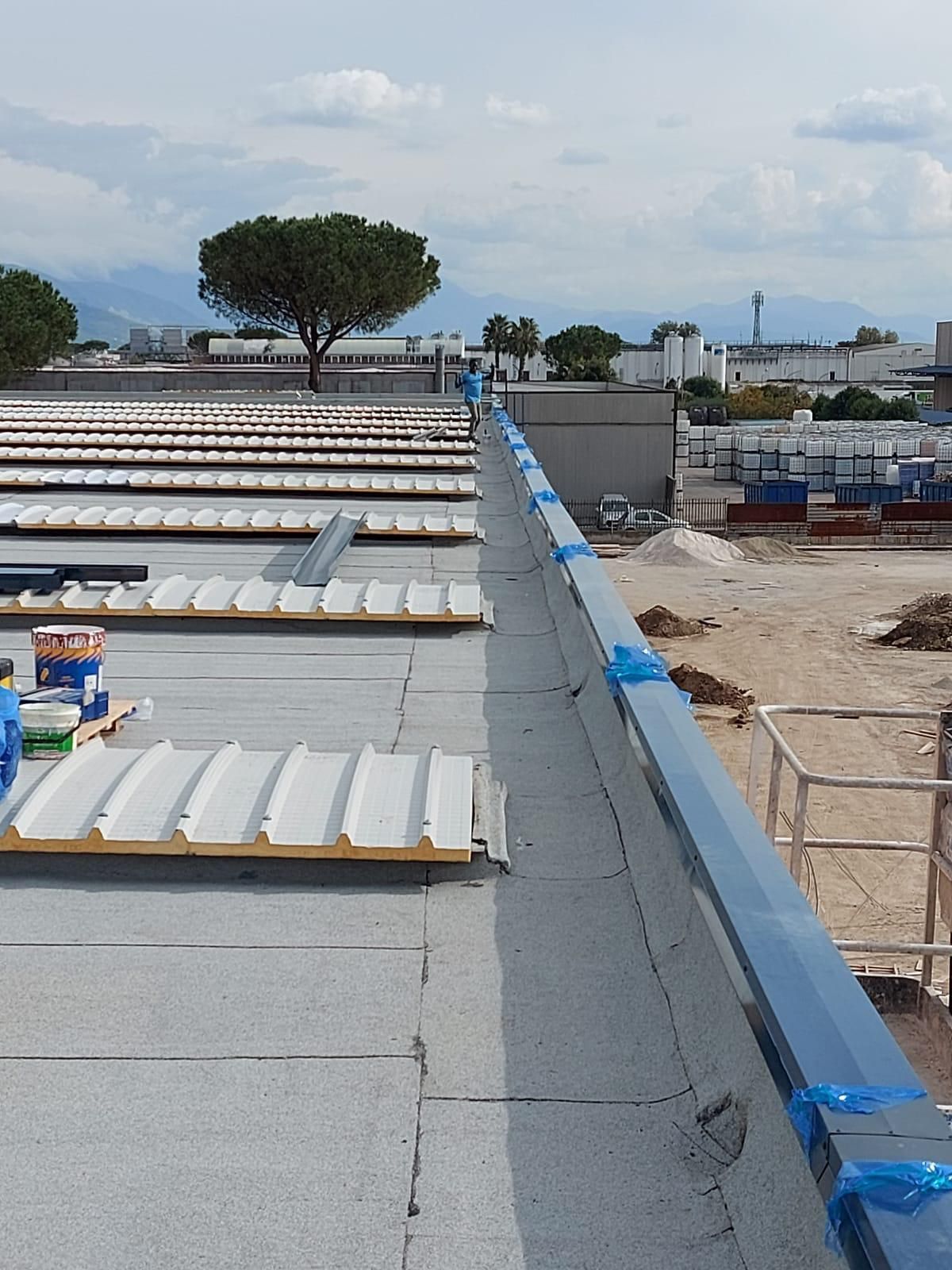Tettoia con pannelli bianchi e bordi blu affacciata su un cantiere e su un lontano orizzonte alberato.