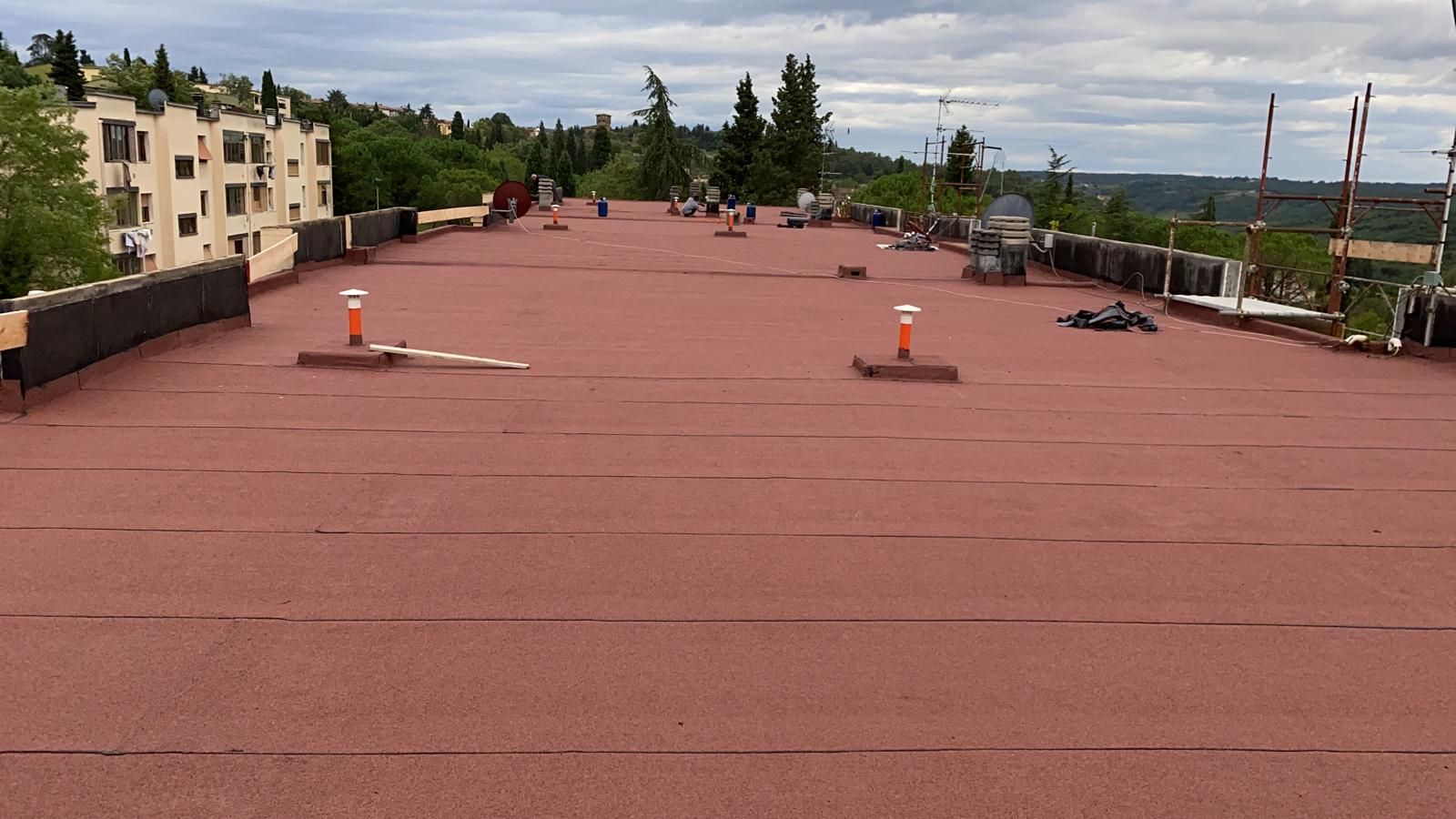 Terrazza sul tetto di colore rosso con prese d'aria sparse, alberi ed edifici lungo i bordi, sotto un cielo nuvoloso.