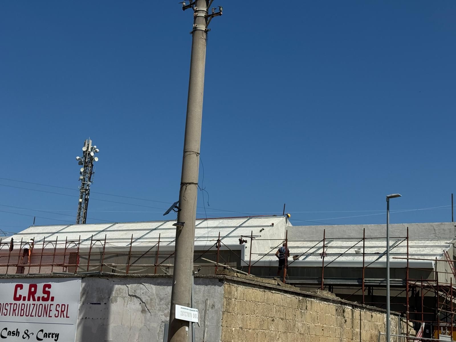 Un edificio dal tetto bianco in costruzione, dietro un muro di cemento e pali della luce, si staglia contro un cielo azzurro.
