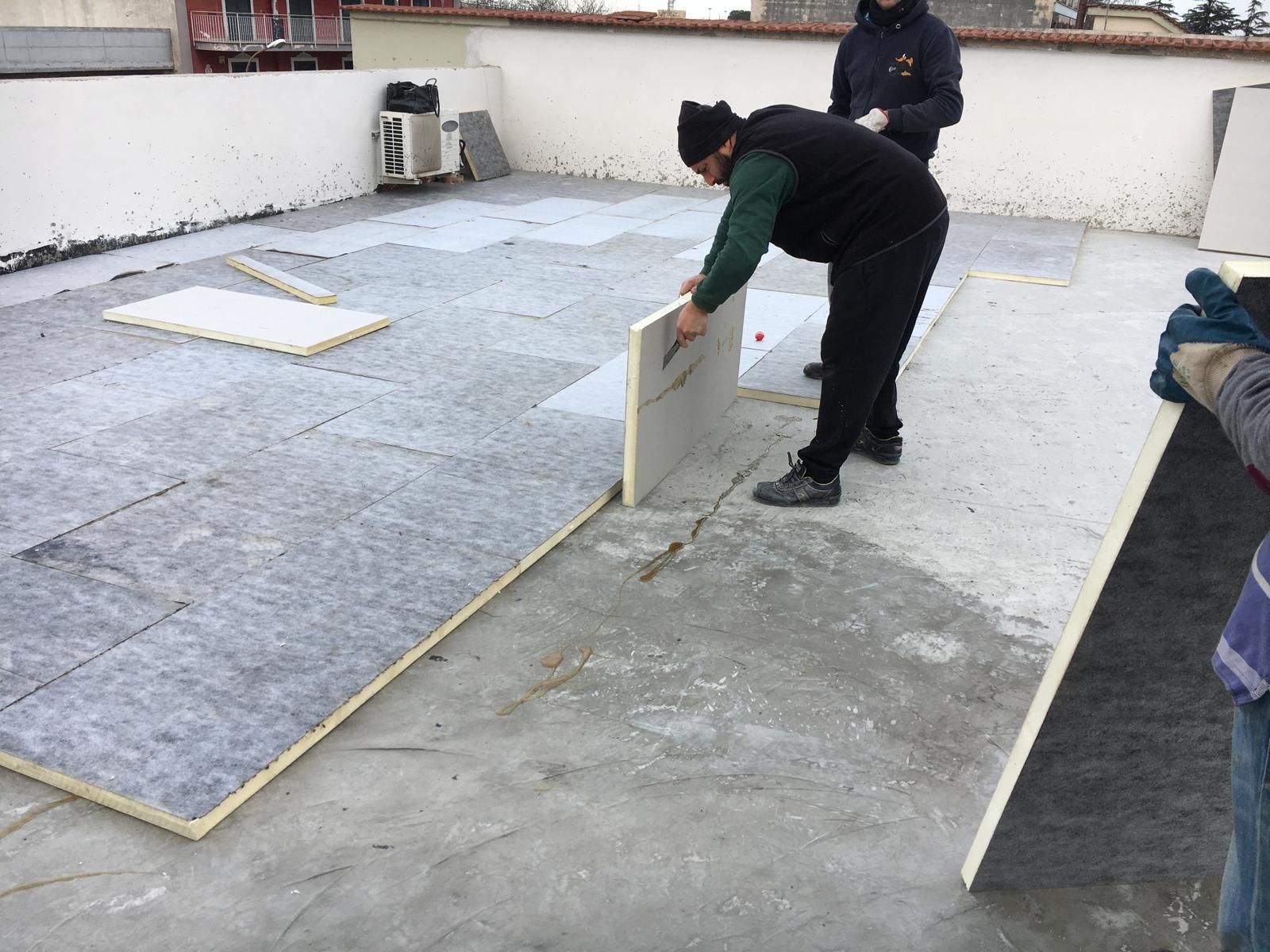 Alcuni operai installano pannelli isolanti su un tetto piano, con una persona china su una tavola grigia.