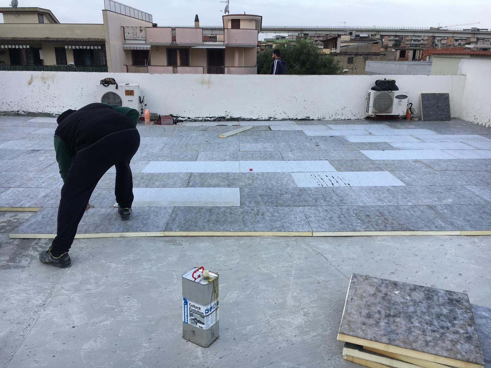 Una persona installa delle tegole su un tetto piano, con attrezzi e materiali nelle vicinanze.