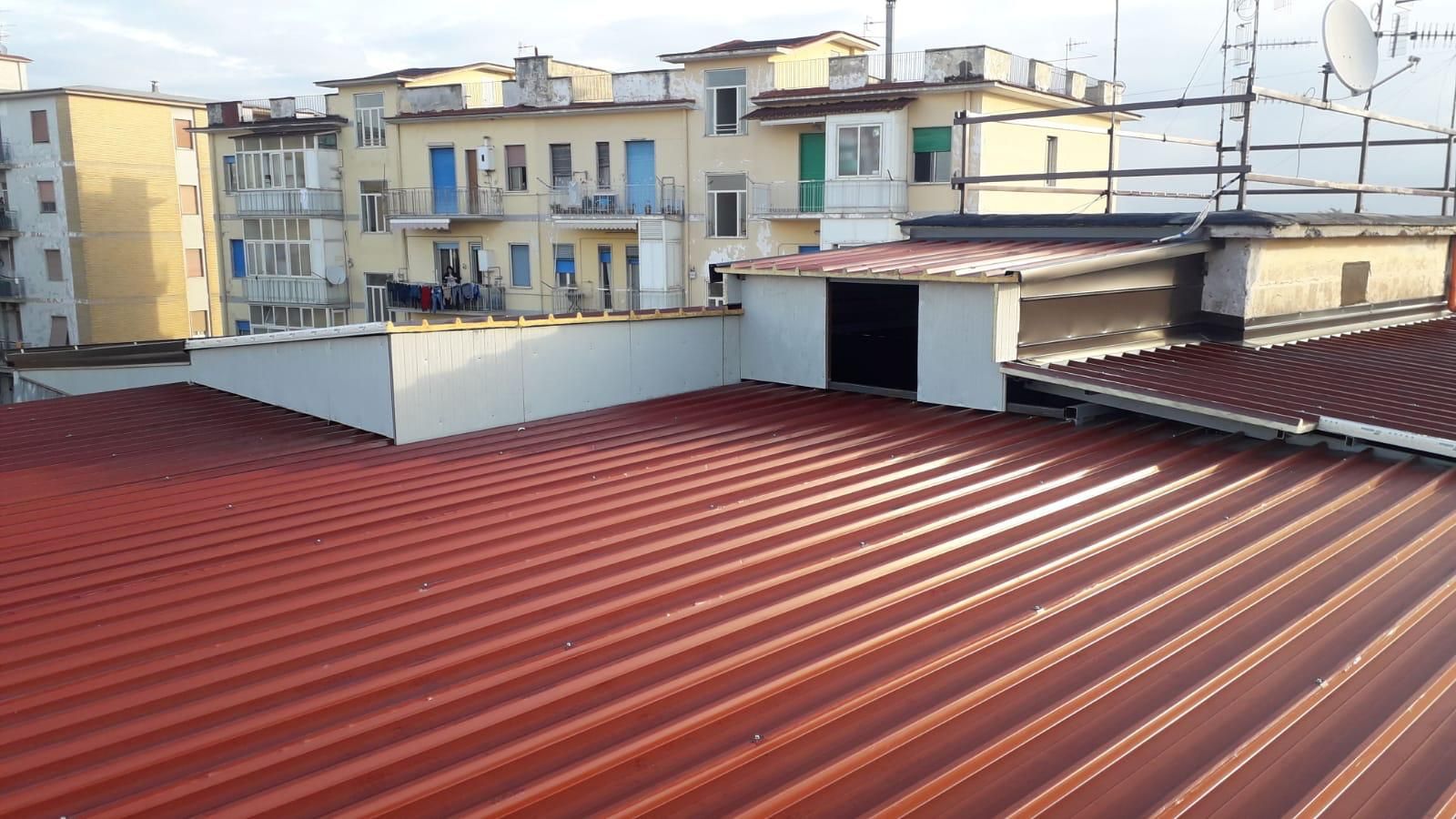 Tettoia con copertura in lamiera ondulata rossa che si affaccia su una fila di palazzine beige sotto un cielo nuvoloso.