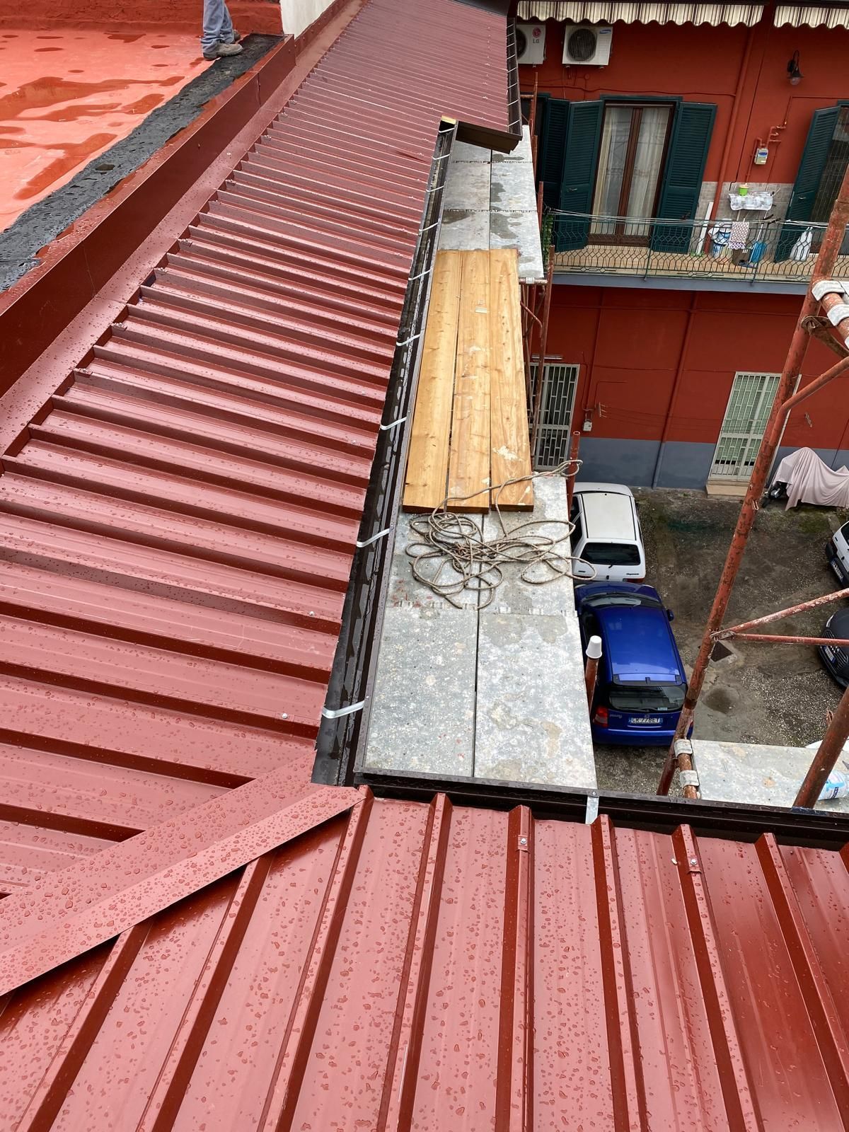 Tetto in metallo rosso con una stretta passerella in assi di legno accanto a un cortile di un edificio rosso