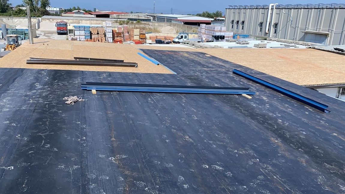 Cantiere edile sul tetto con membrana impermeabilizzante nera e travi metalliche blu posate lungo la copertura.