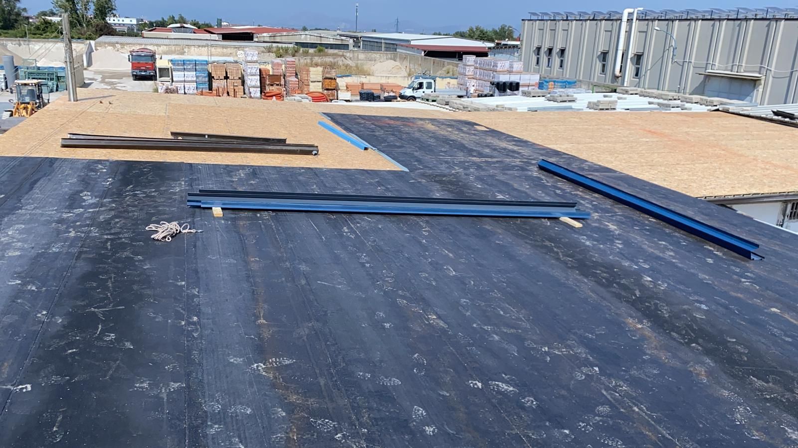 Cantiere edile sul tetto con membrana impermeabilizzante nera e travi metalliche blu posate lungo la copertura.