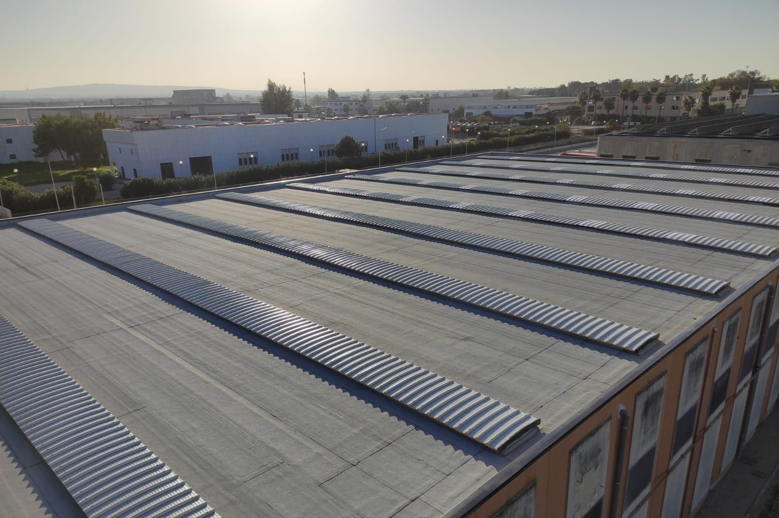 Veduta aerea di un grande tetto industriale con file di lucernari e degli edifici circostanti al tramonto.
