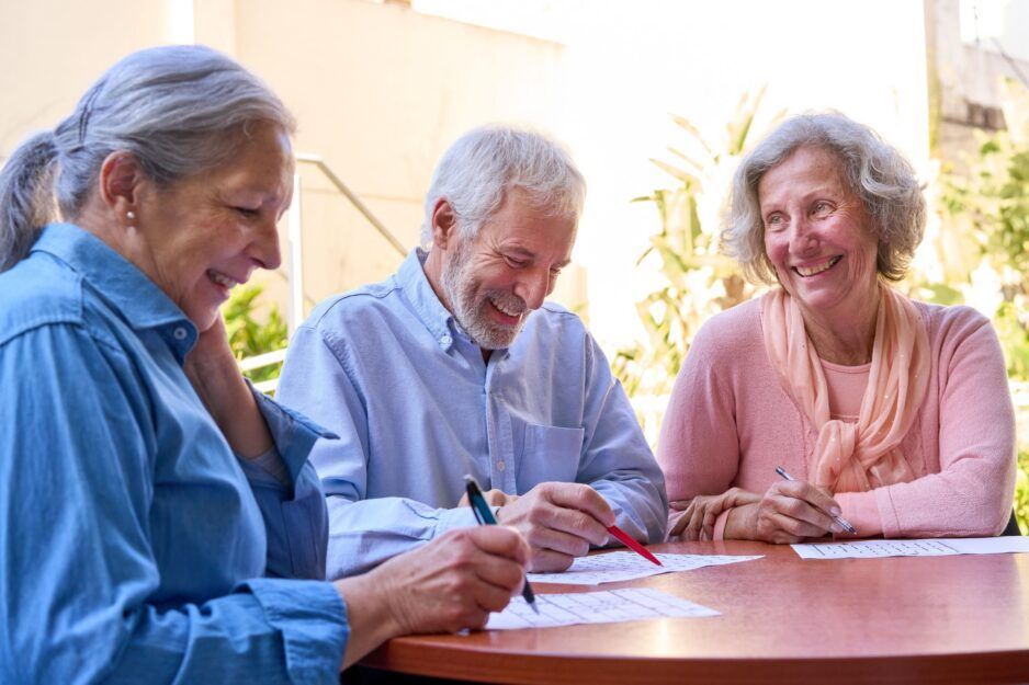 Three people sit around a round table, smiling and working together on papers with pens in a bright, outdoor setting.