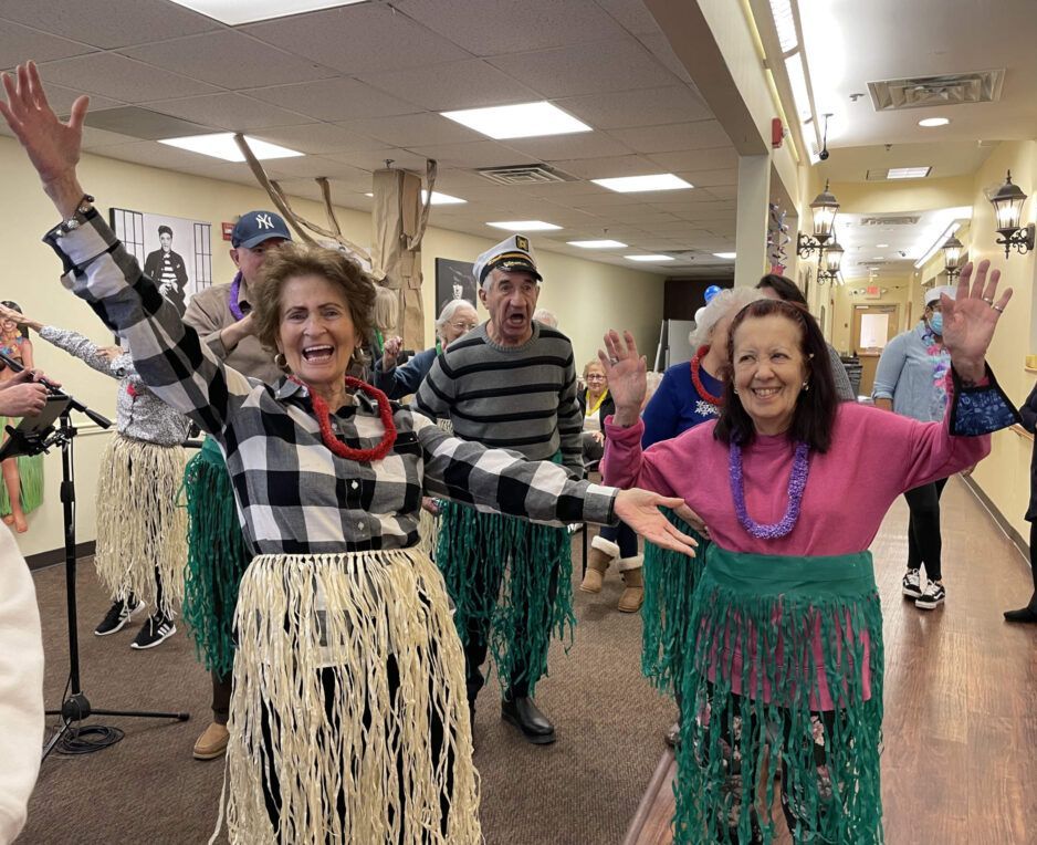 People in hula-style skirts and leis pose with arms raised in a hallway, smiling during an indoor activity.