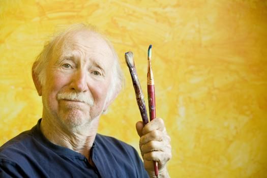 An artist holds two paintbrushes in front of a textured yellow background.